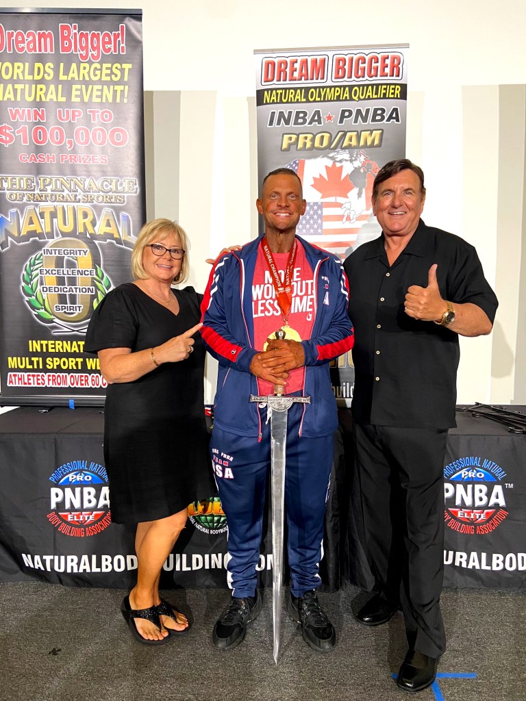 A tanned bodybuilder holds a sword surrounded by two happy acquaintances.
