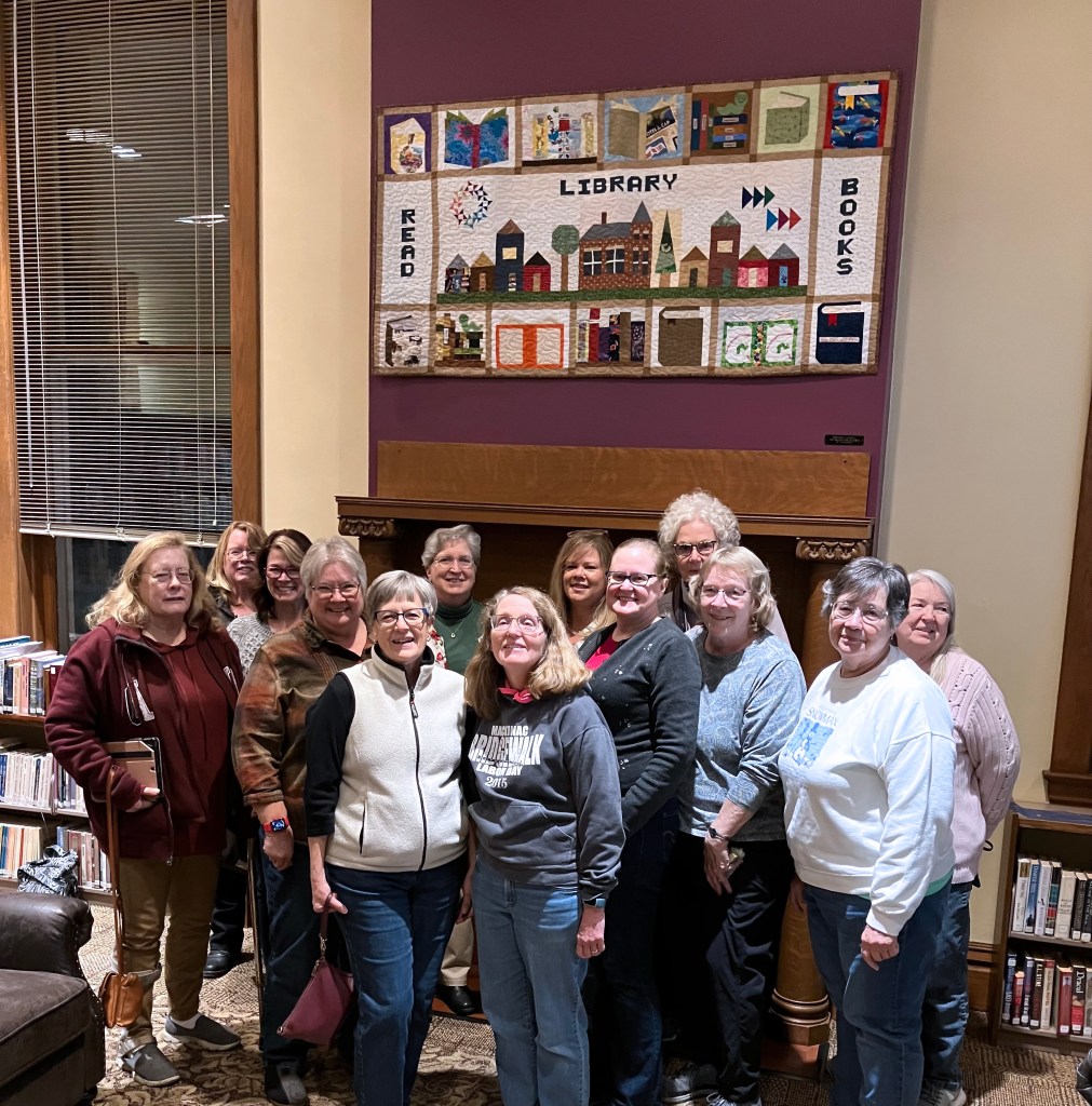 A group of women stands in front of a library-themed quilt.