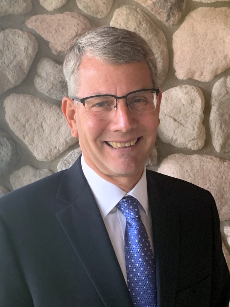 A man with glasses wearing a suit stands in front of a fieldstone wall.