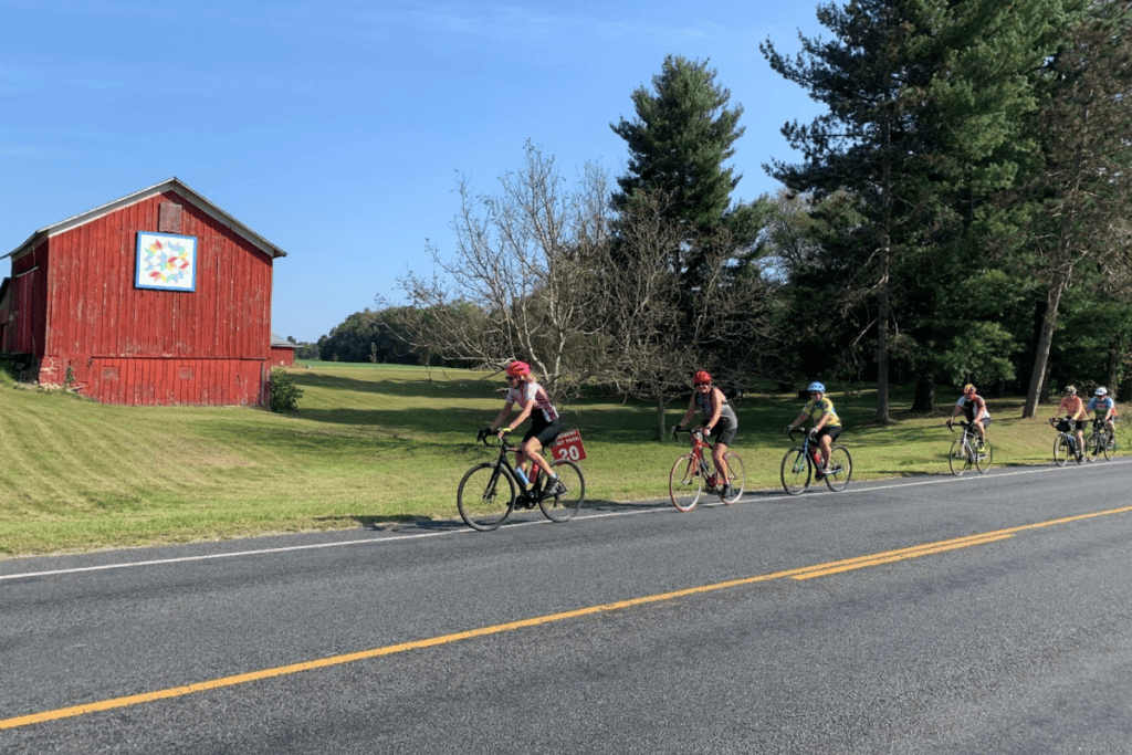 Bikers go along a country road with a red barn in the background.
