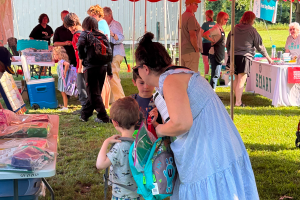 A woman in a blue dress stands with two young kids wearing backpacks looking at tables set up under a tent.