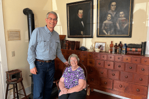 A man and a woman (standing and seated) stand in a historic home with painted portraits on the wall.