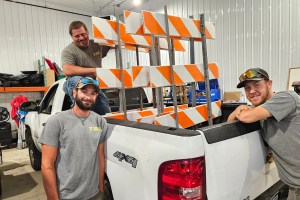 Three men stand near a truck holding caution signs that are used to block roads.