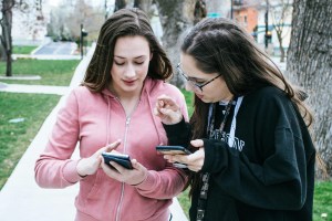 Two teenage girls look at cell phones while outdoors.