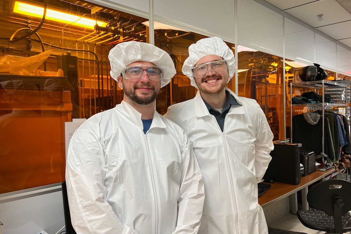 pauli1 Two men in safety goggles and white protective suits stand in a science lab.