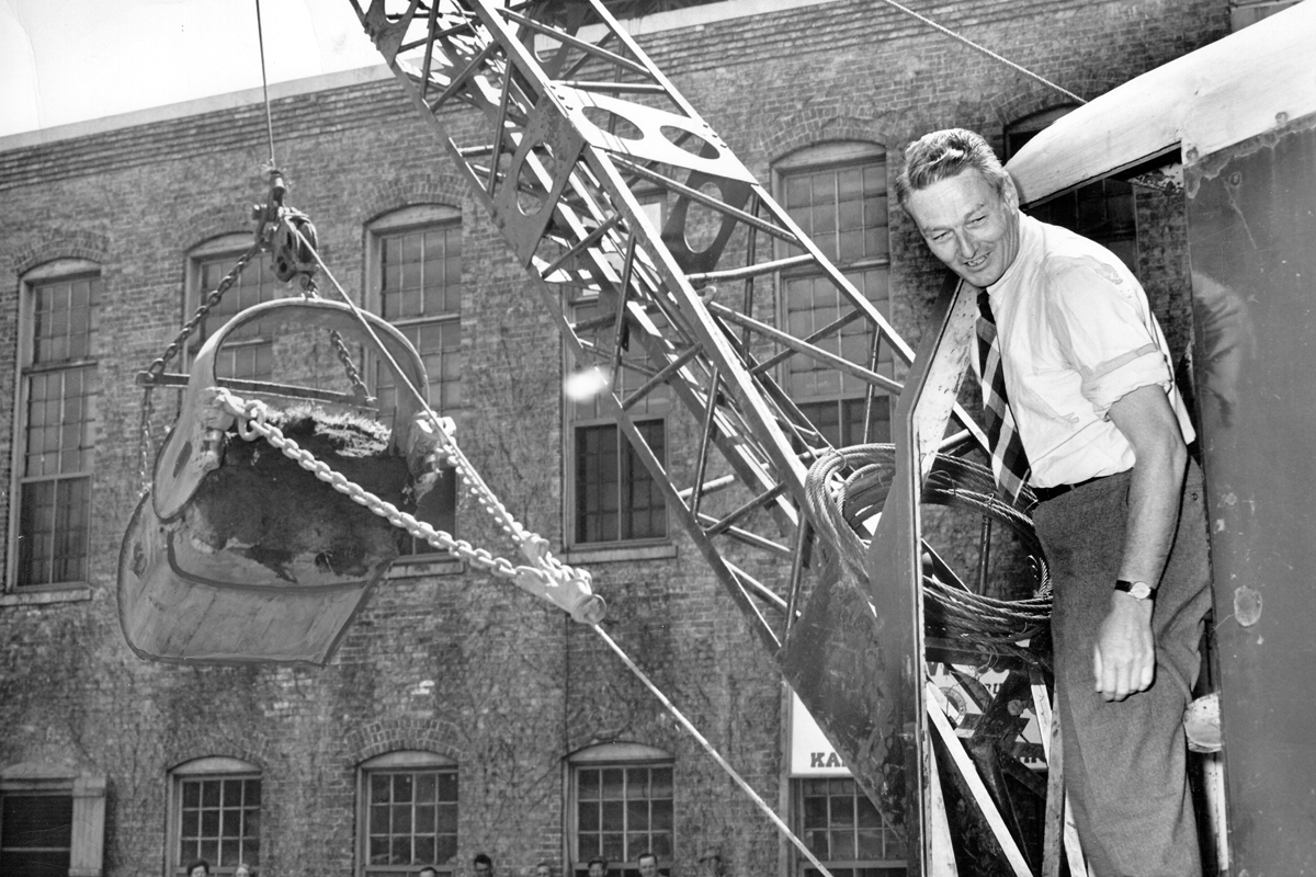 A man operates a large crane by a brick building in an old photograph.