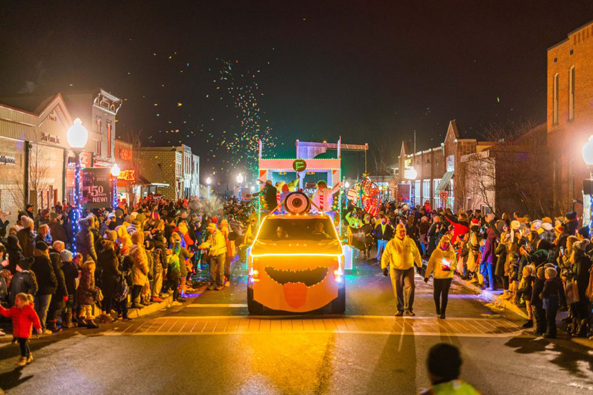 A festive vehicle comes down the street surrounded by lights and onlookers in heavy coats.