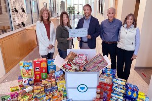 Five individuals pose with a large check and food donation boxes.