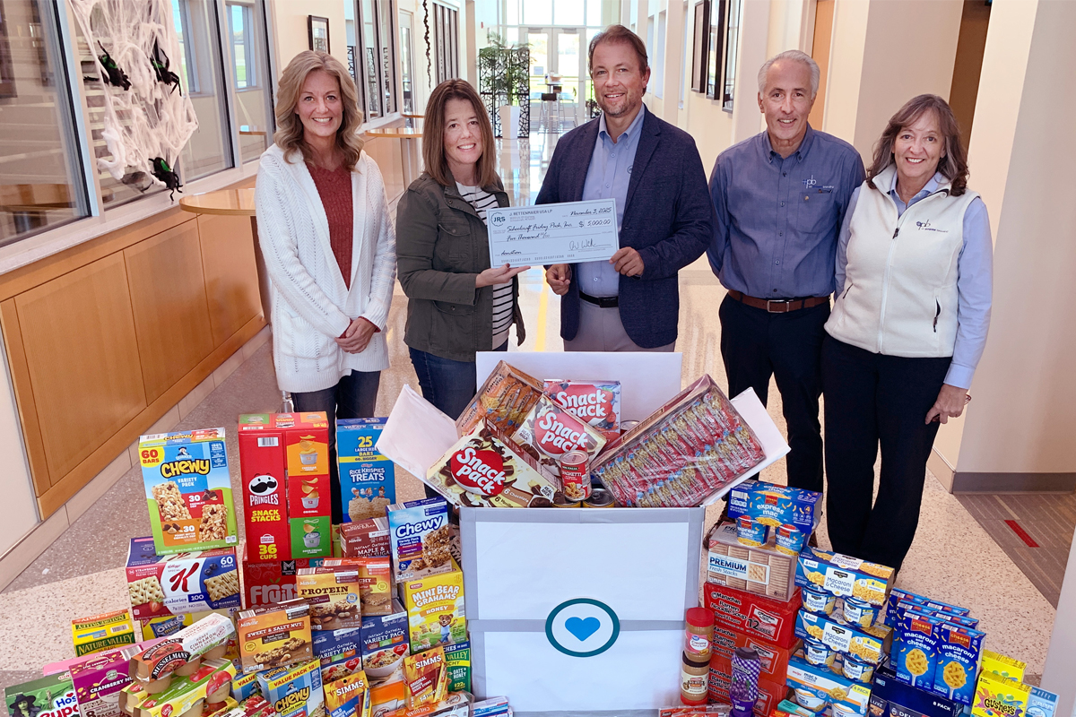 Five individuals pose with a large check and food donation boxes.