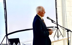A white-haired man stands at a microphone.