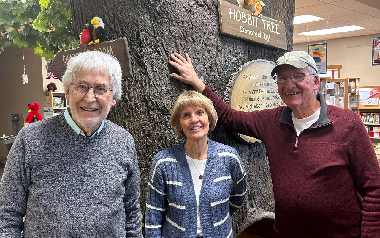 Three people stand near a fake tree inside a library building.