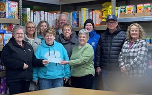A woman hands a check to a group standing in a food pantry with packaged goods filling the shelves.