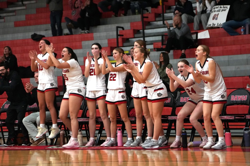 Several girls in white basketball uniforms cheer from the sidelines.