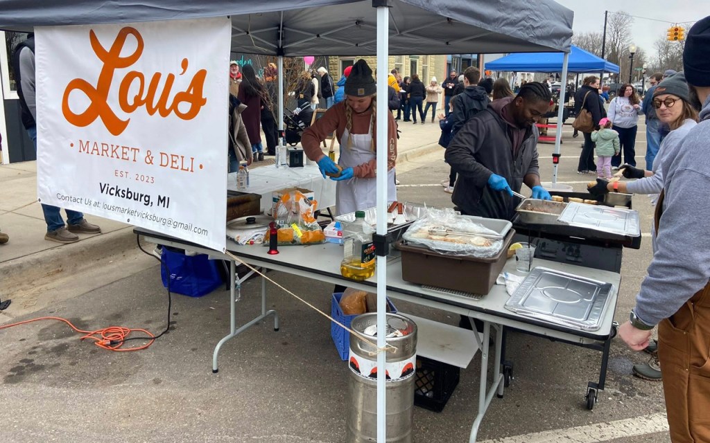 A chef makes hamburger sliders at a tent with a sign that reads Lou's Market & Deli.