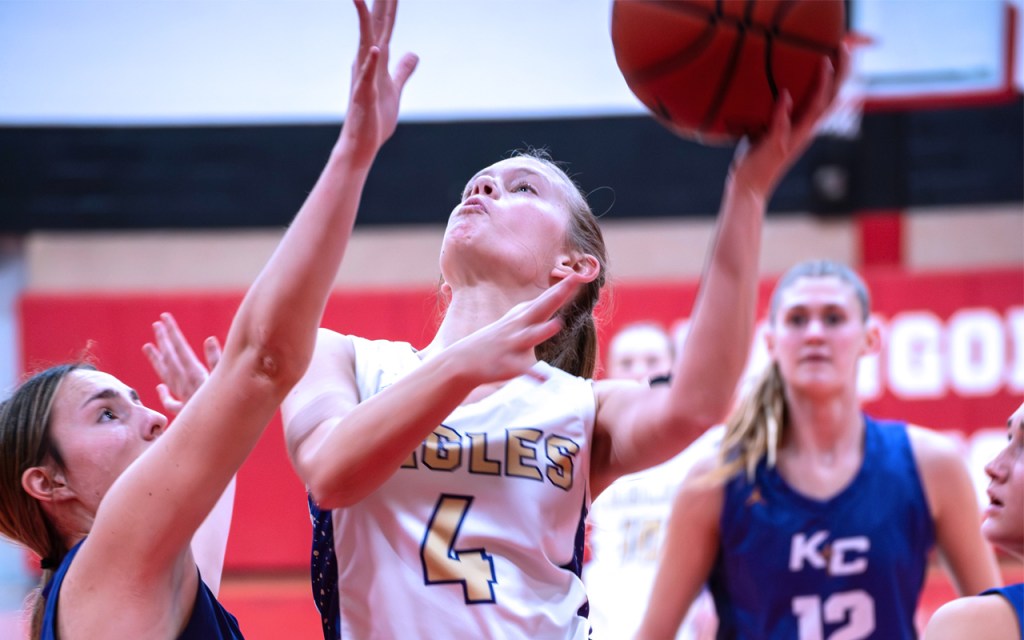 A basketball player leaps up with the ball while the other team's player tries to block the shot.