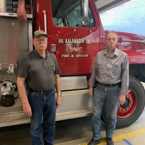 Two men stand in front of a red fire truck.