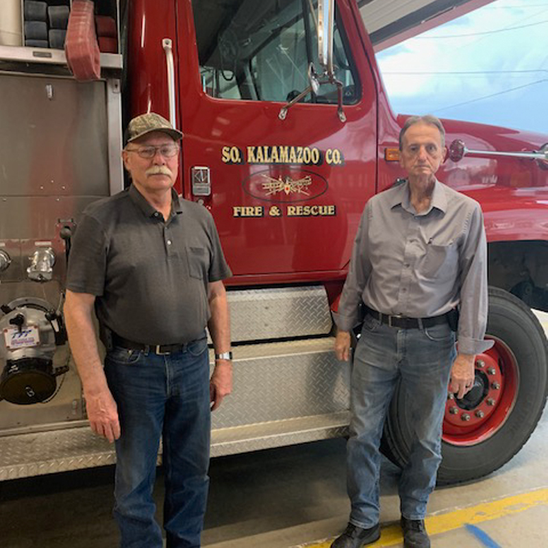 Two men stand in front of a red fire truck.