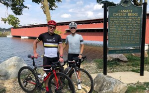 Two men in helmets stand with their bikes by the water and a covered bridge.