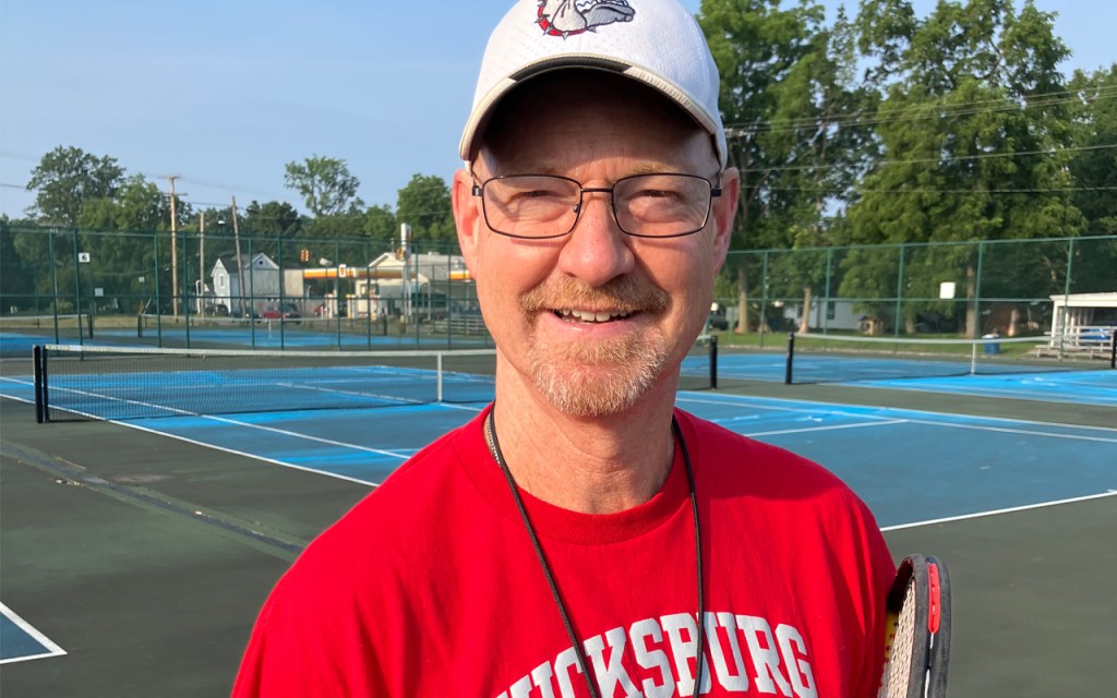 A man in a red shirt with a whistle and a cap, holds a tennis racket.