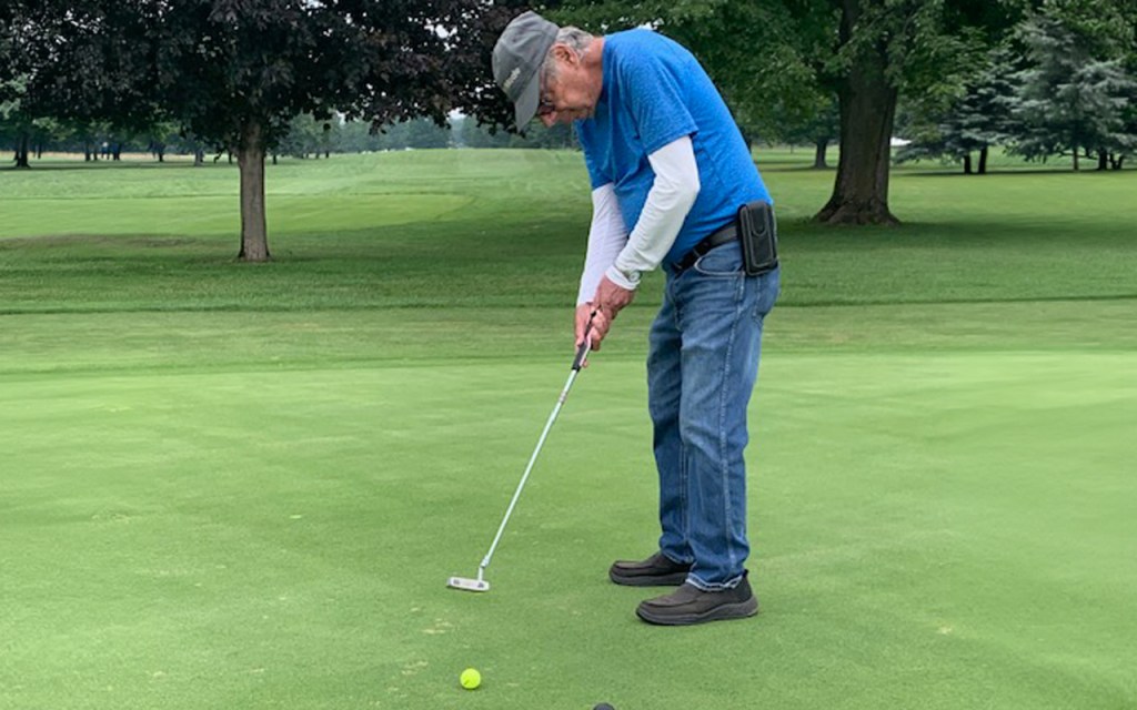 A man in a blue shirt and jeans looks down to line up a golf swing.